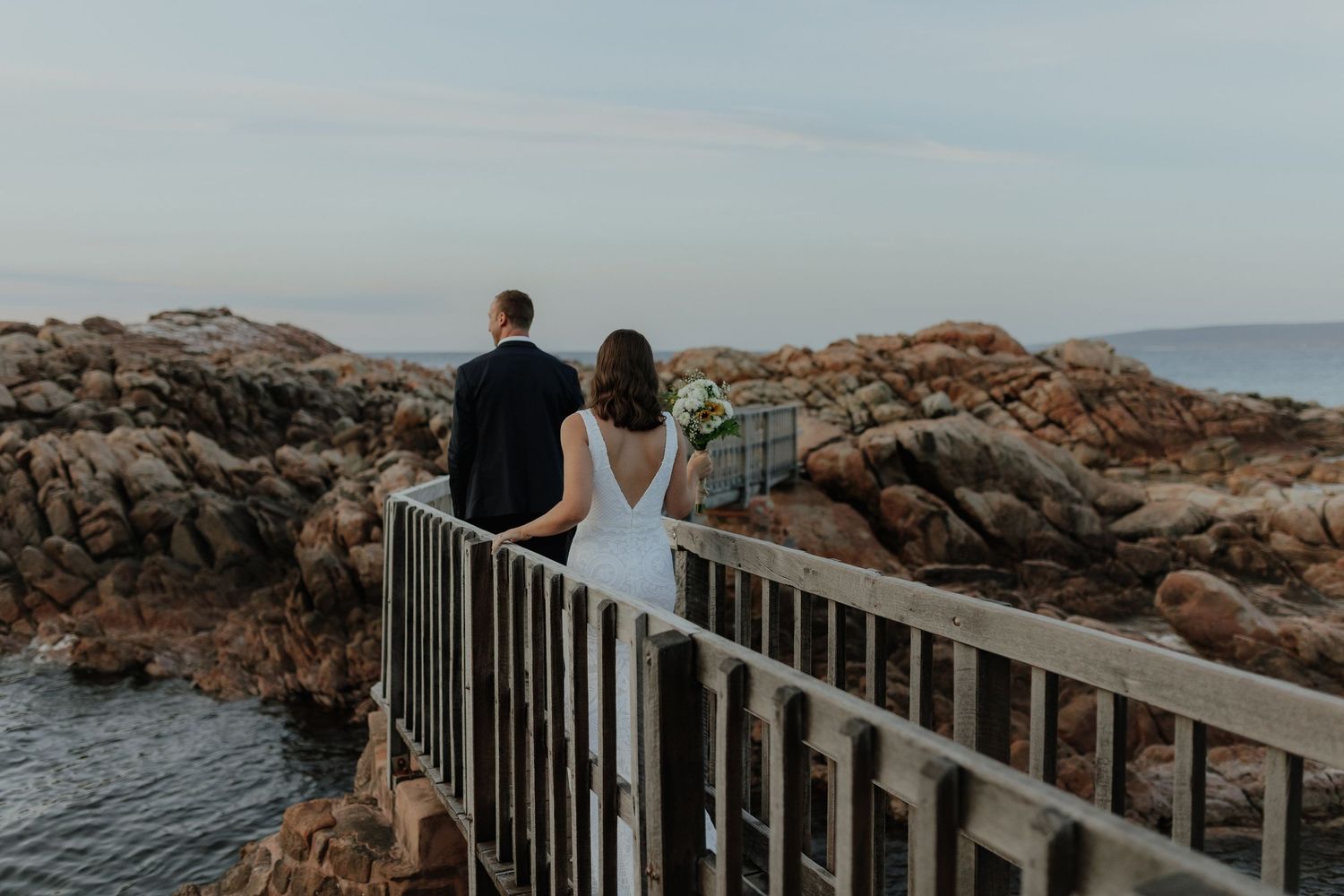Wedding couple standing on a metal bridge overlooking rocky coastal waters at dusk.