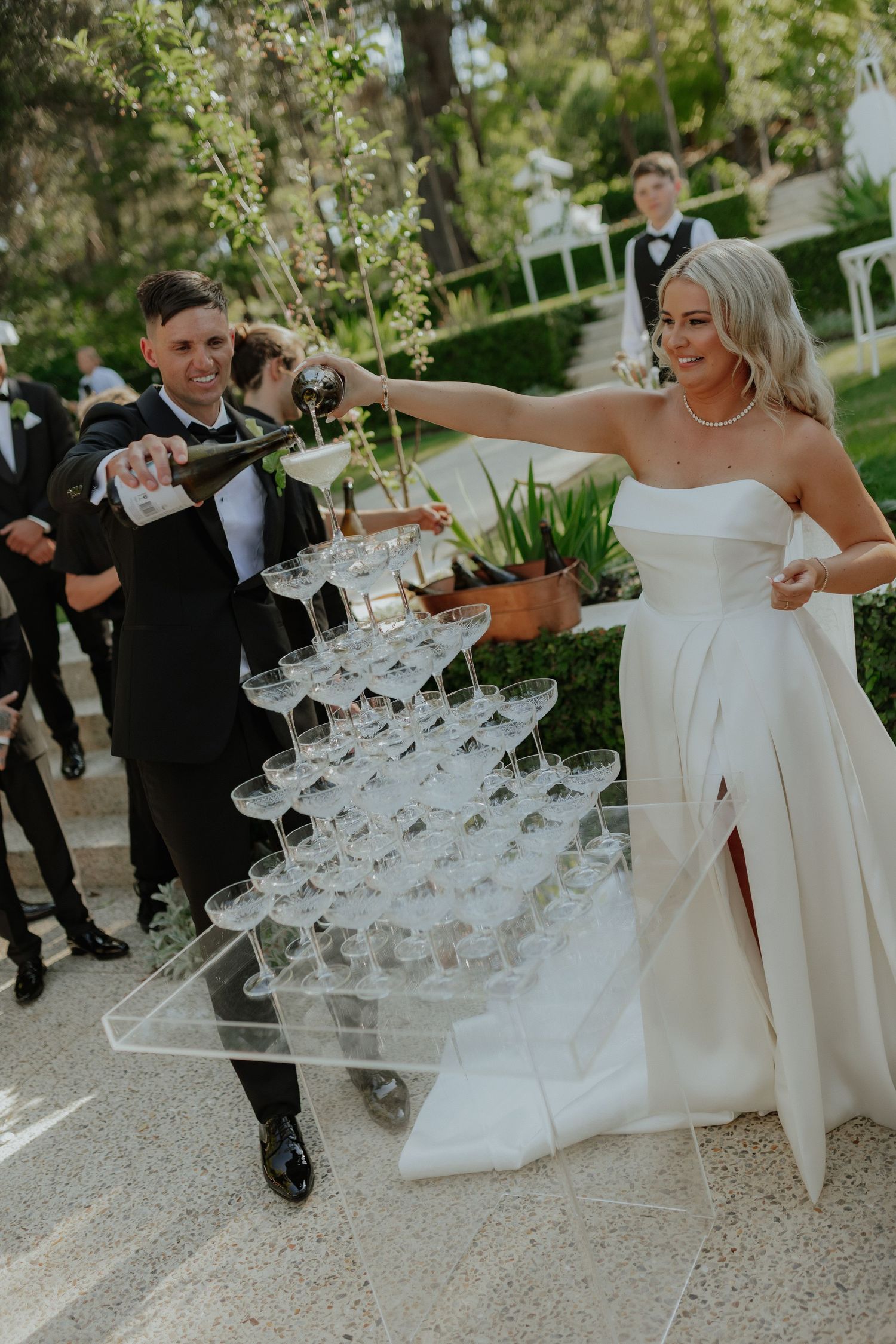 Bride and groom pour champagne tower at elegant outdoor wedding reception surrounded by guests.