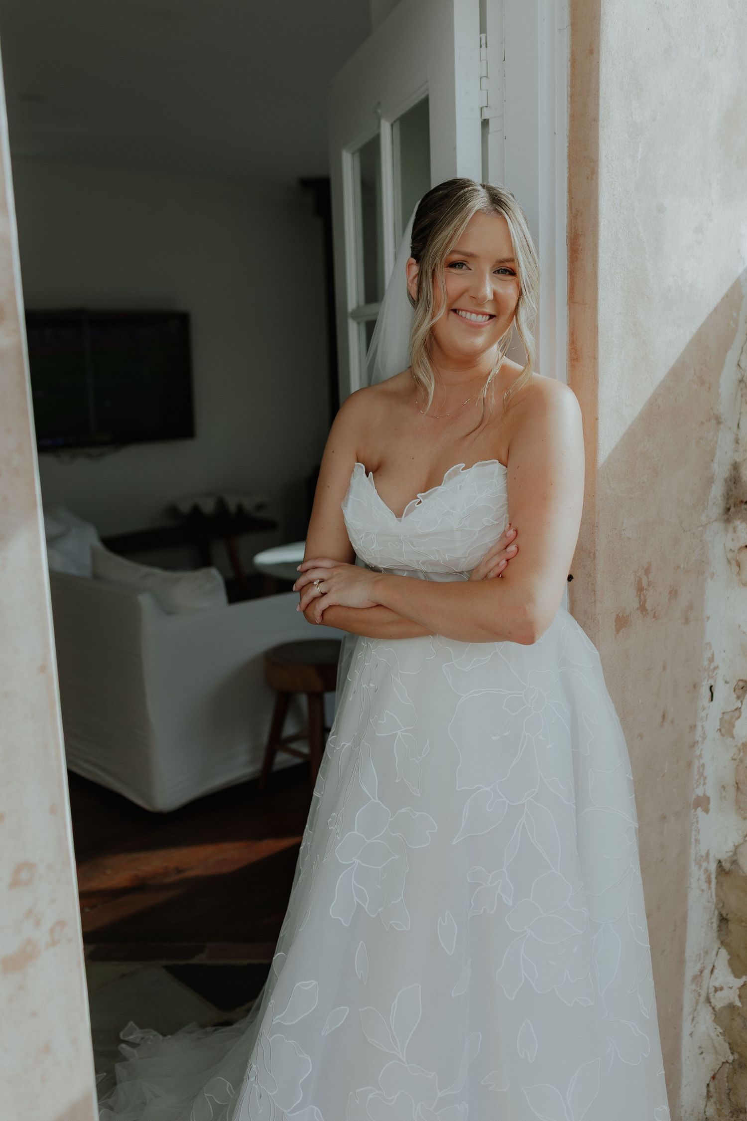 Smiling bride in strapless white floral wedding gown standing in a doorway with arms crossed.
