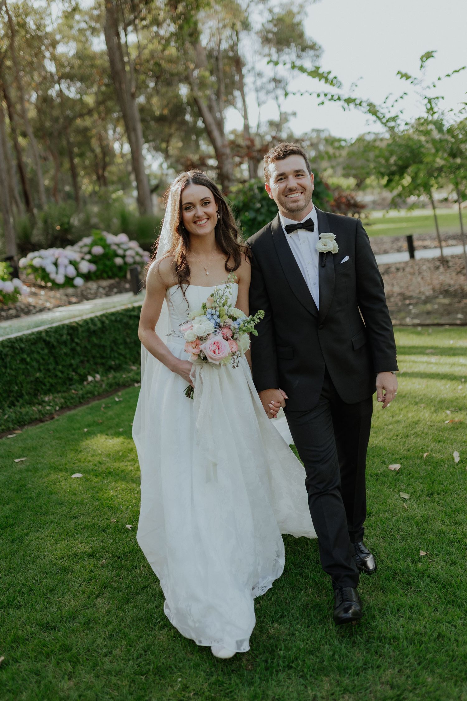 Happy bride and groom walking hand in hand through a lush garden, bride holding floral bouquet.