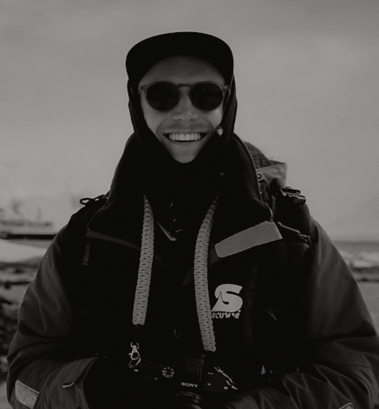 Smiling photographer in black jacket and cap wearing sunglasses at waterfront, black and white photo.