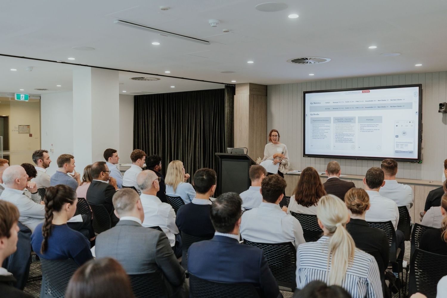 Business presenter at podium addressing a full room of seated professionals at a corporate conference presentation.