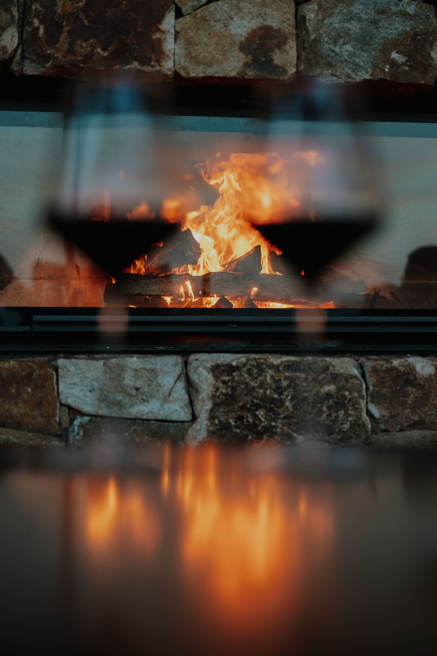 Two glasses of red wine silhouetted against a glowing stone fireplace with warm flames reflecting on a surface.