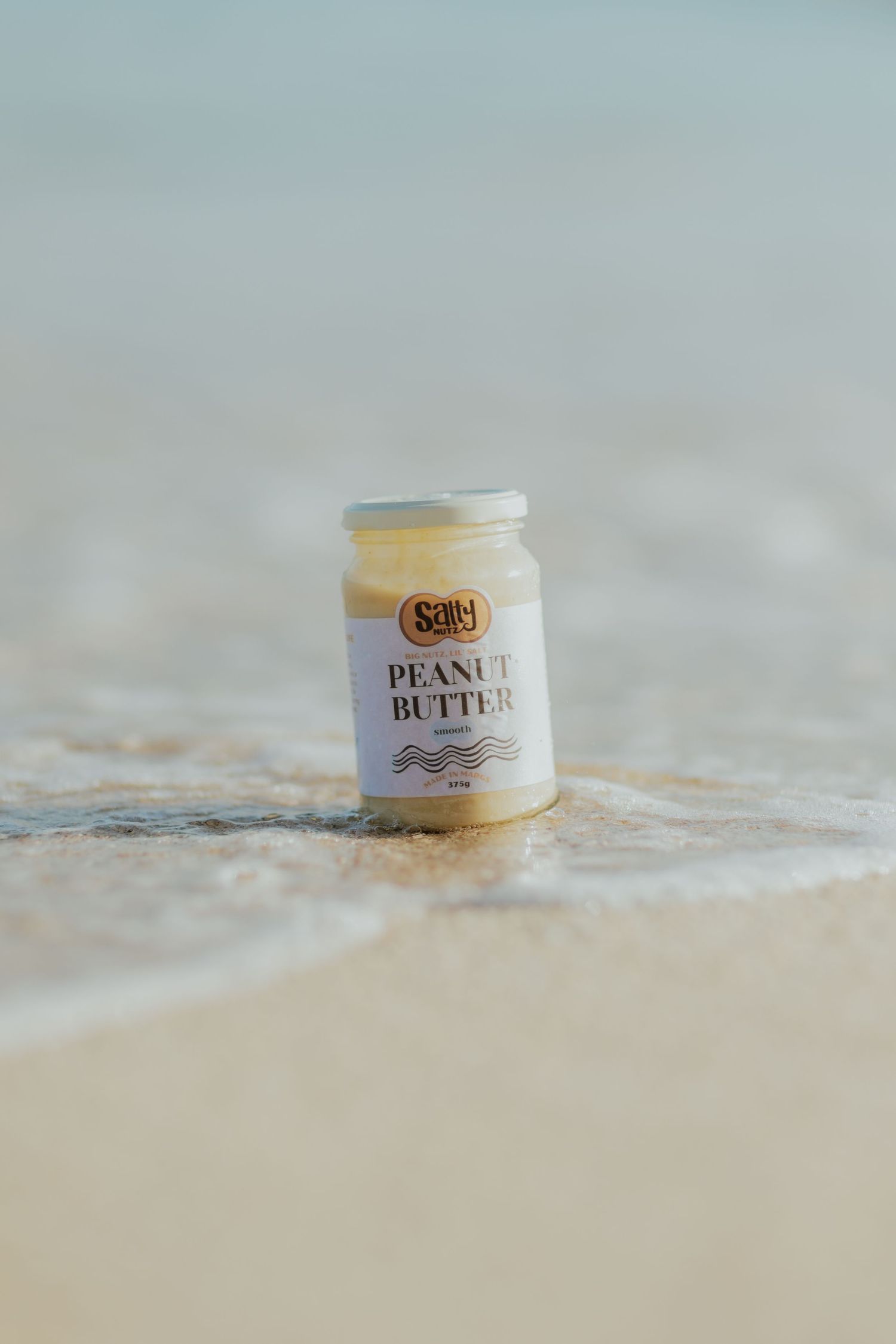 A jar of Sally peanut butter sits on a sandy beach with ocean waves in the background.