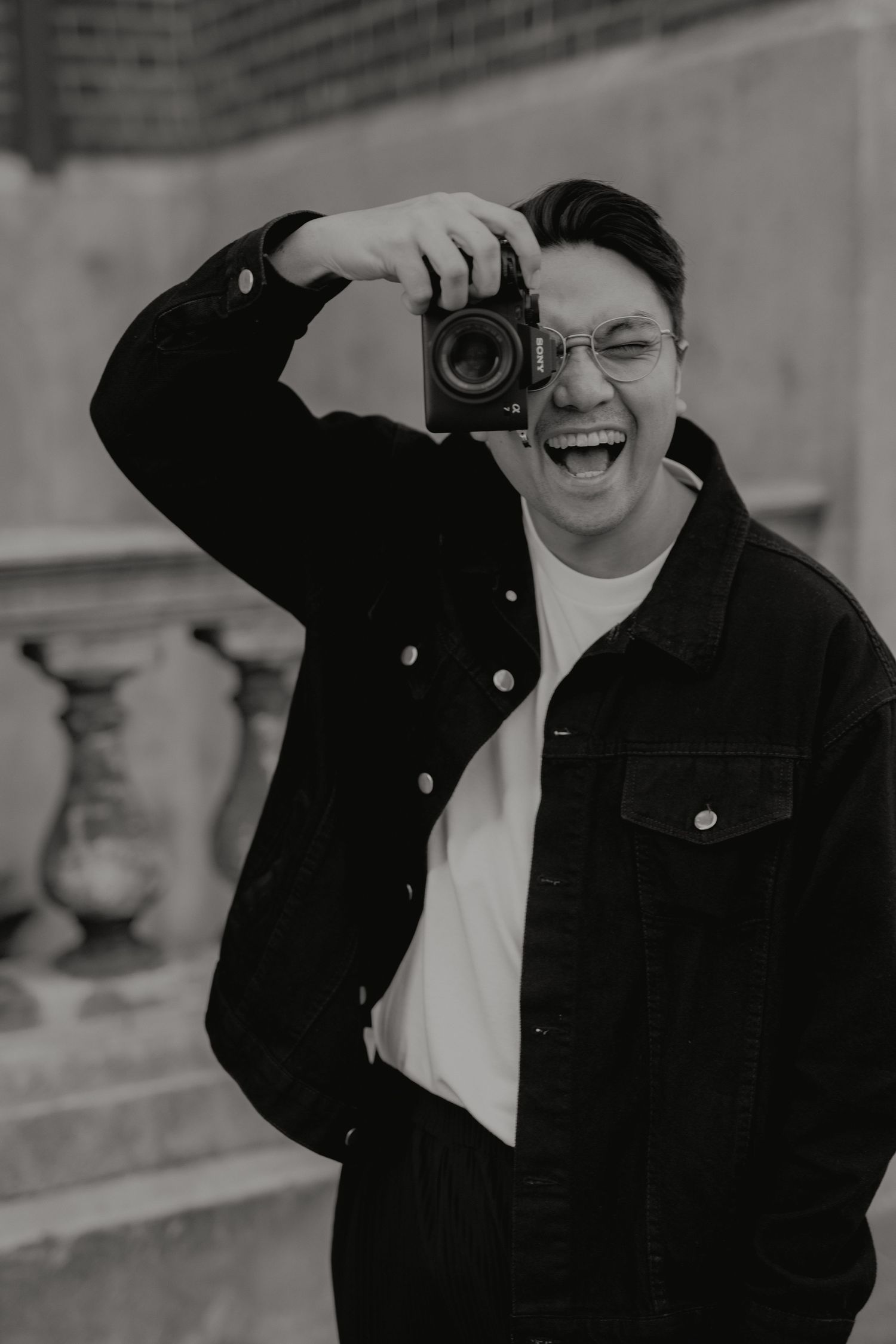 Laughing man in black denim jacket holding camera up to face in black and white photo.