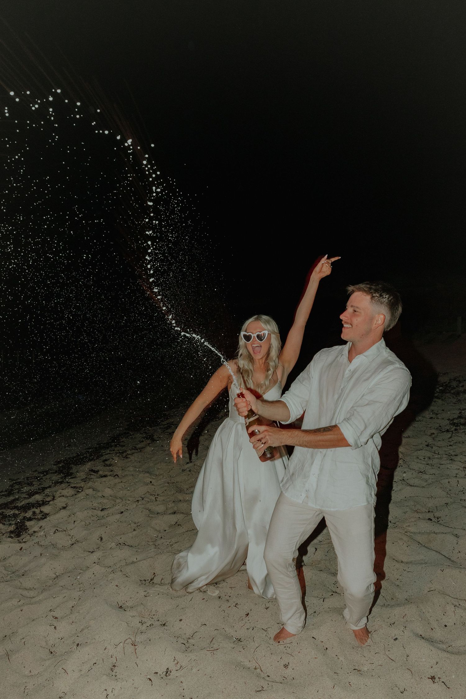 Bride and groom celebrate at night on the beach spraying champagne under dark sky.