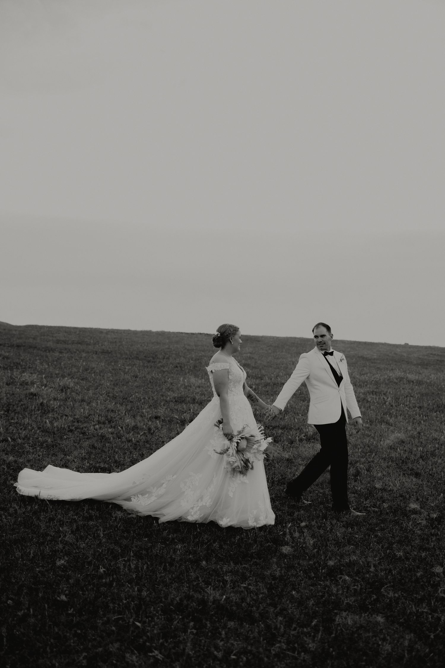 Black and white photo of bride and groom holding hands walking through an open field.