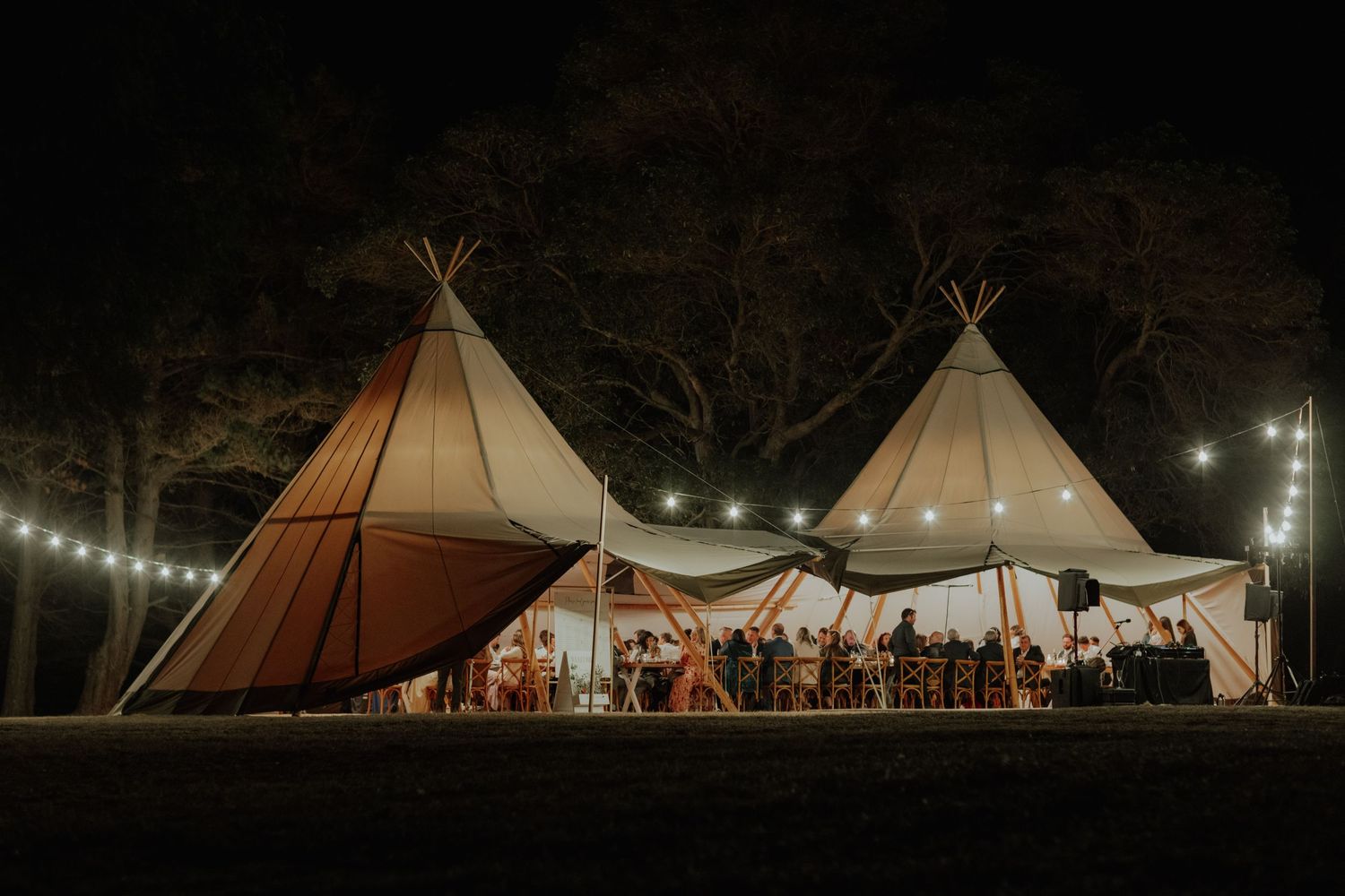 Stunning nighttime wedding reception under two illuminated tipis with festoon lights and guests gathered.