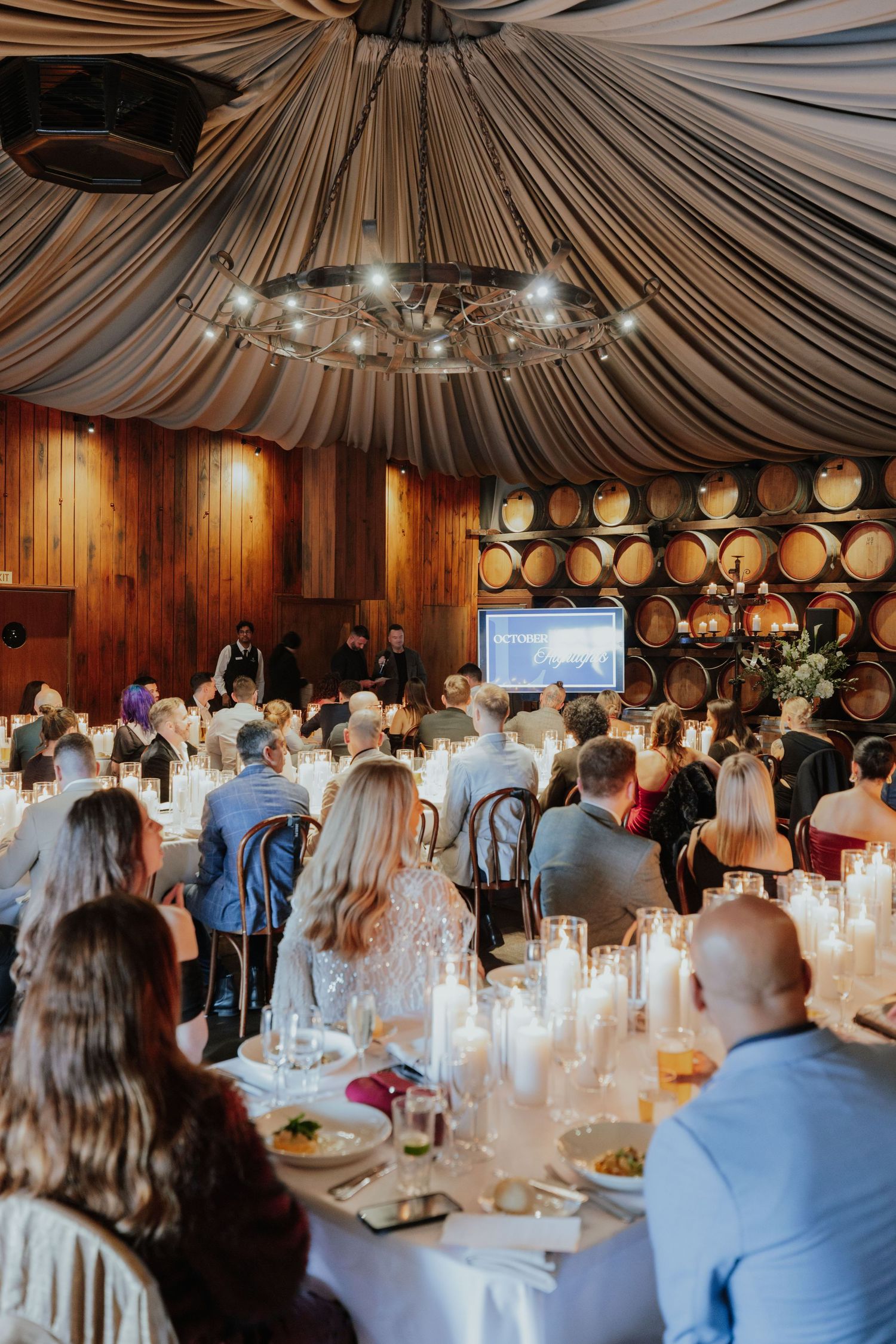 Elegantly dressed guests seated at candlelit tables during a formal winery barrel room gala.
