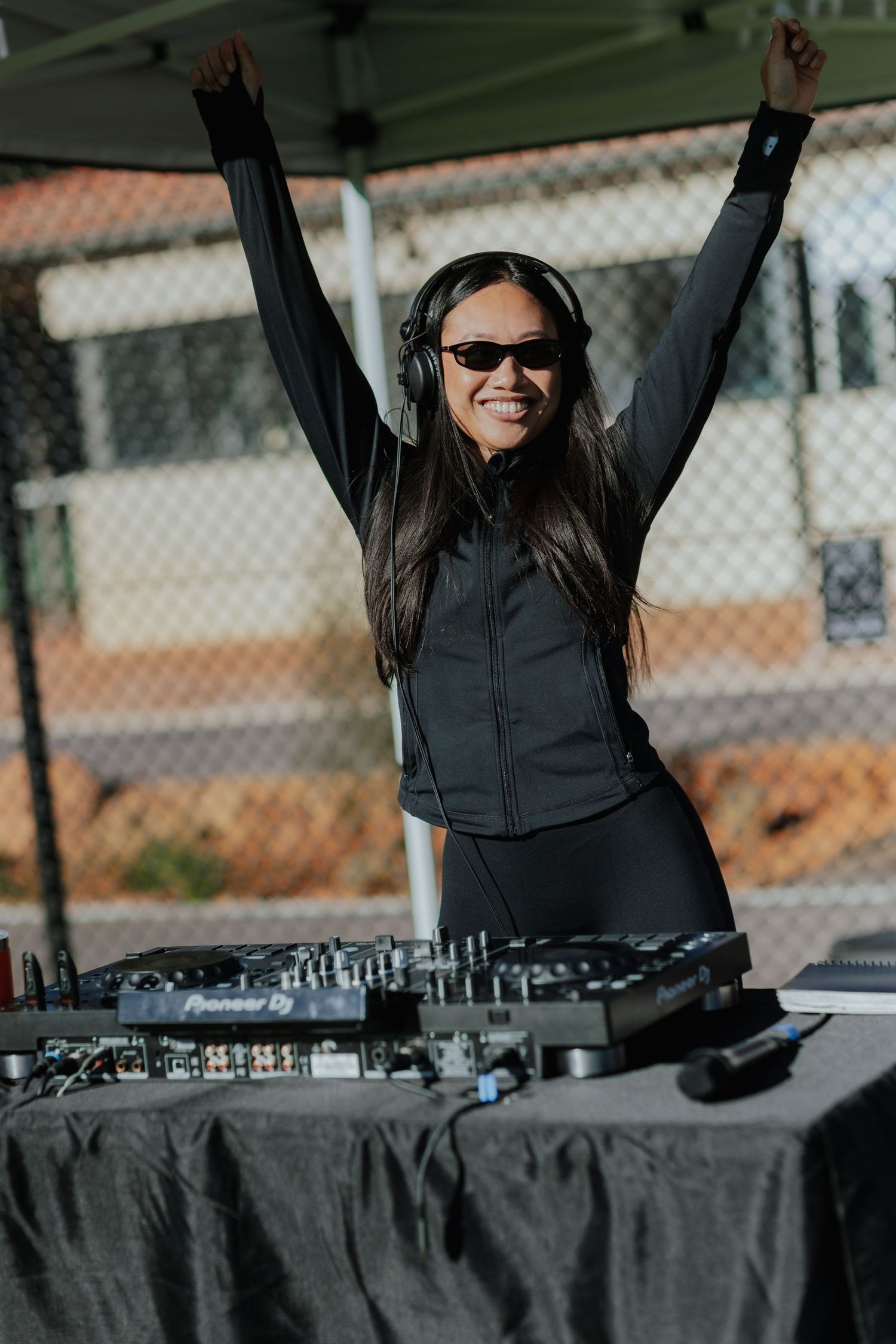 Female DJ with arms raised celebrating behind Pioneer DJ decks at an outdoor event.