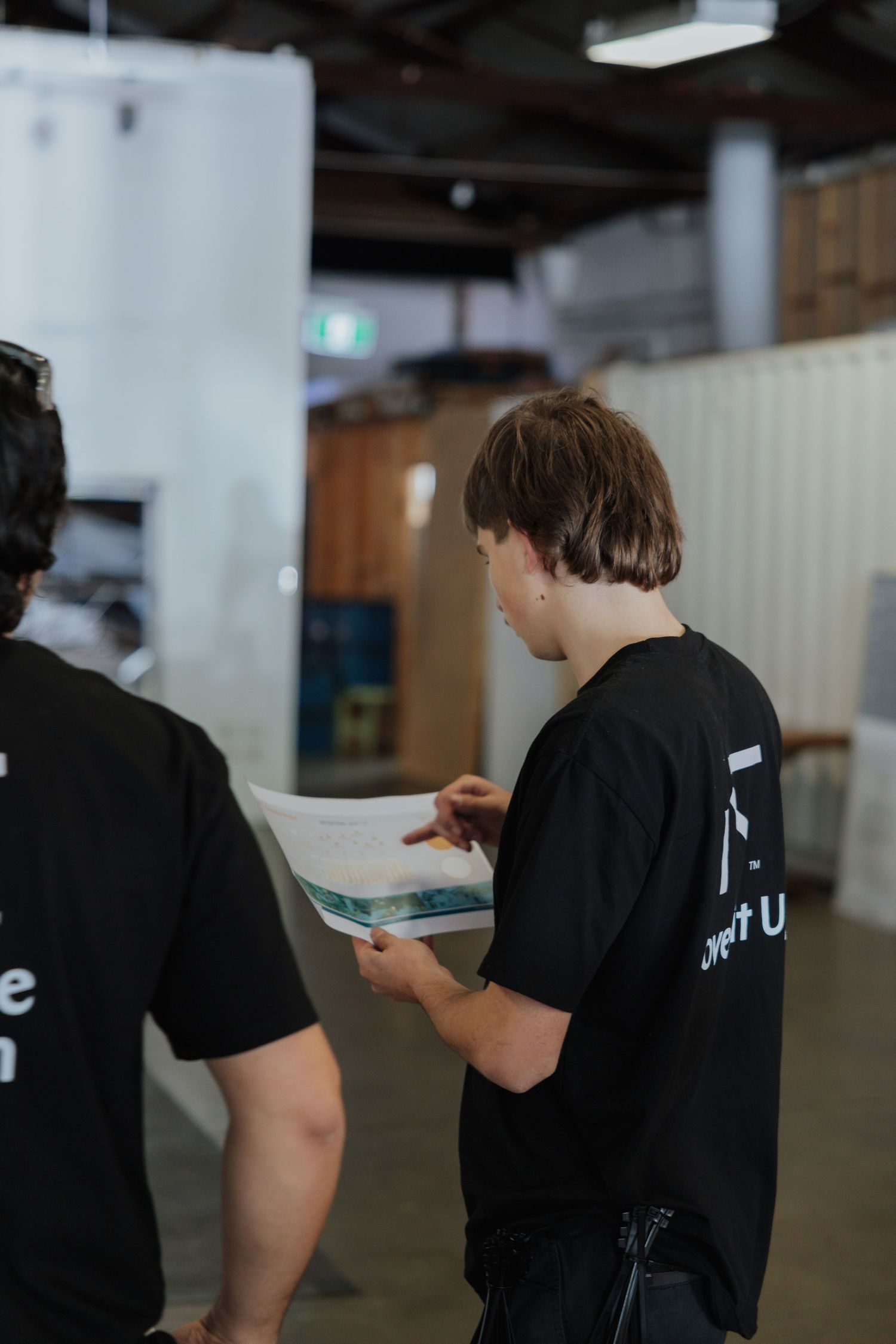 Two workers in black t-shirts reviewing a printed document inside an industrial warehouse space.