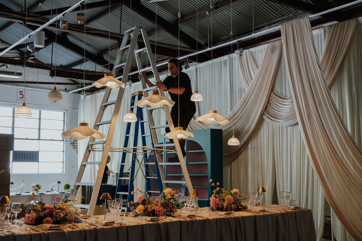 Event worker on a ladder arranging rattan pendant lights above a floral-decorated reception table.