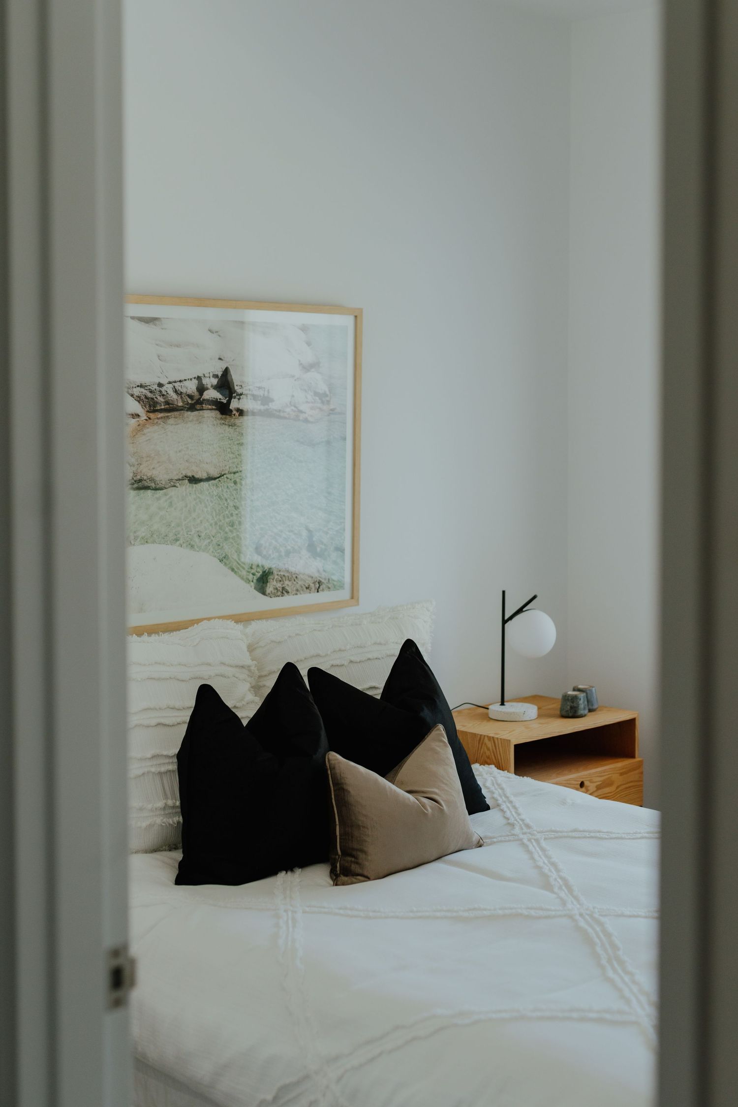 Minimalist bedroom viewed through doorway with framed coastal art, black pillows, and white bedding.