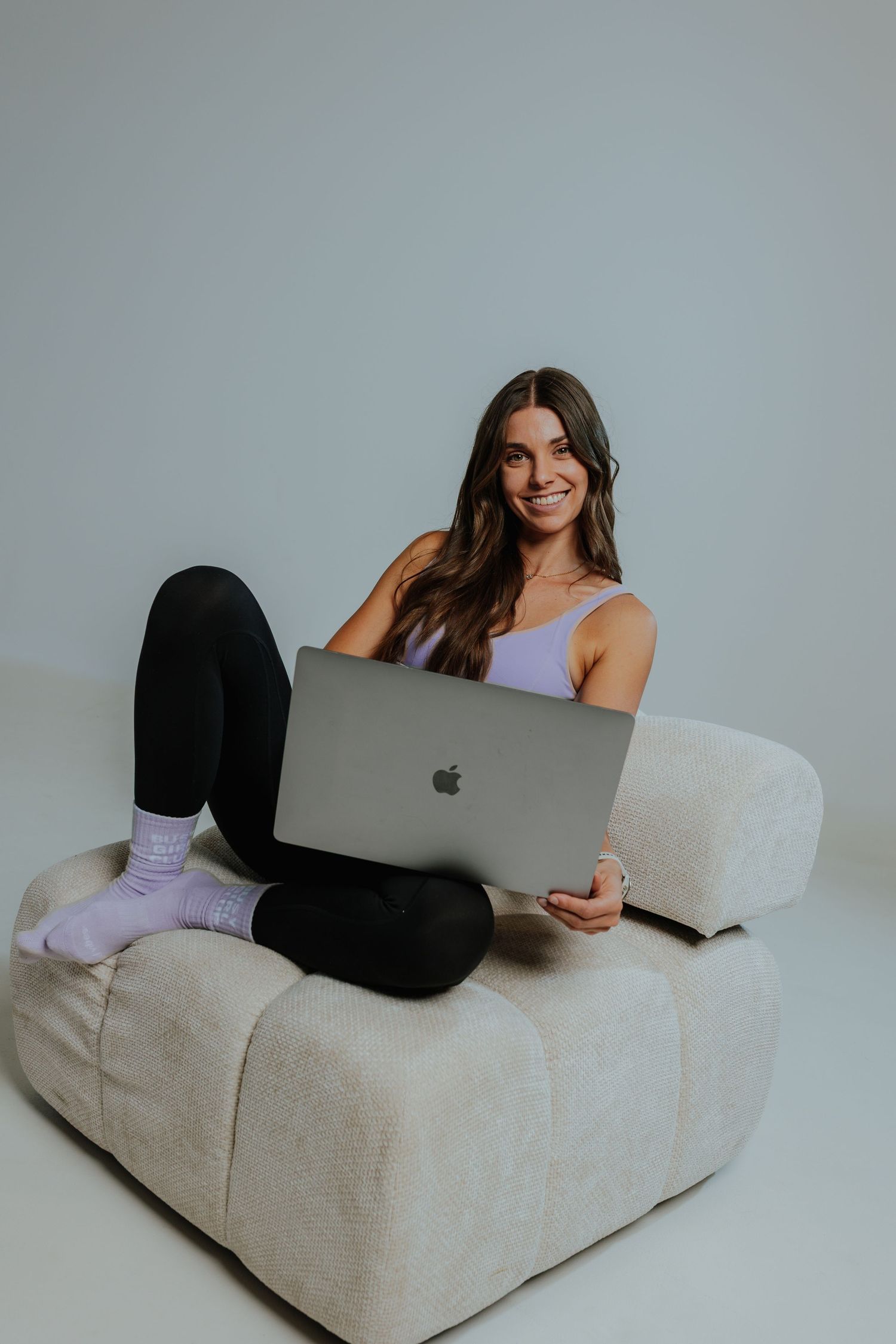 Smiling woman in lavender top sitting on cream sofa using an Apple MacBook laptop.