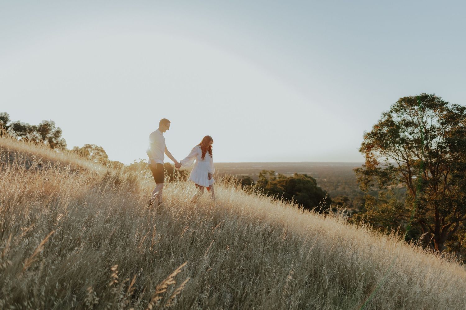 Couple holding hands walking through golden grass fields at sunset.