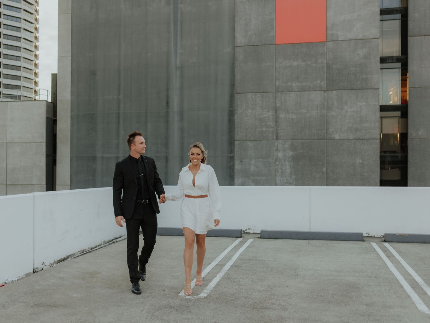 Stylish couple walking hand in hand on rooftop parking lot with urban backdrop.
