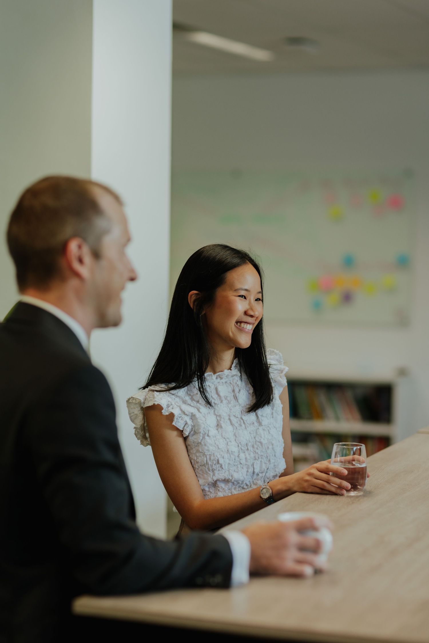 Smiling Asian woman in white lace top conversing with a colleague at a wooden table in an office.