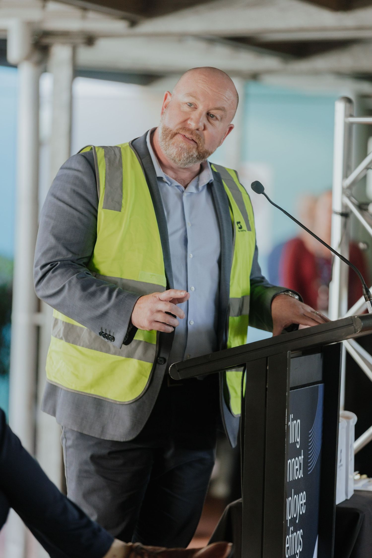 Man in yellow safety vest speaking at a podium during an outdoor corporate event.