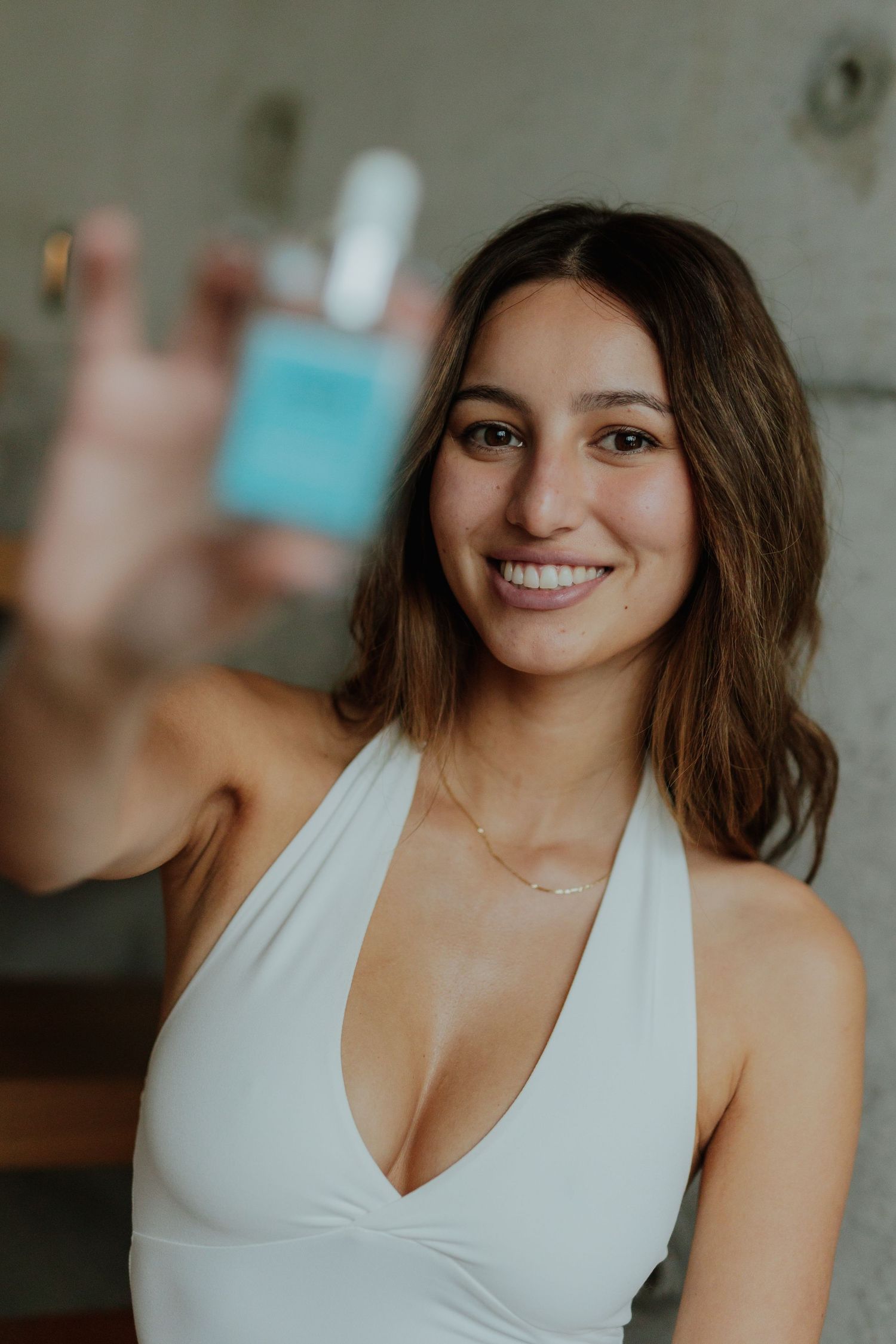 Smiling brunette woman in white halter top holding up a teal Nomii skincare product toward camera.