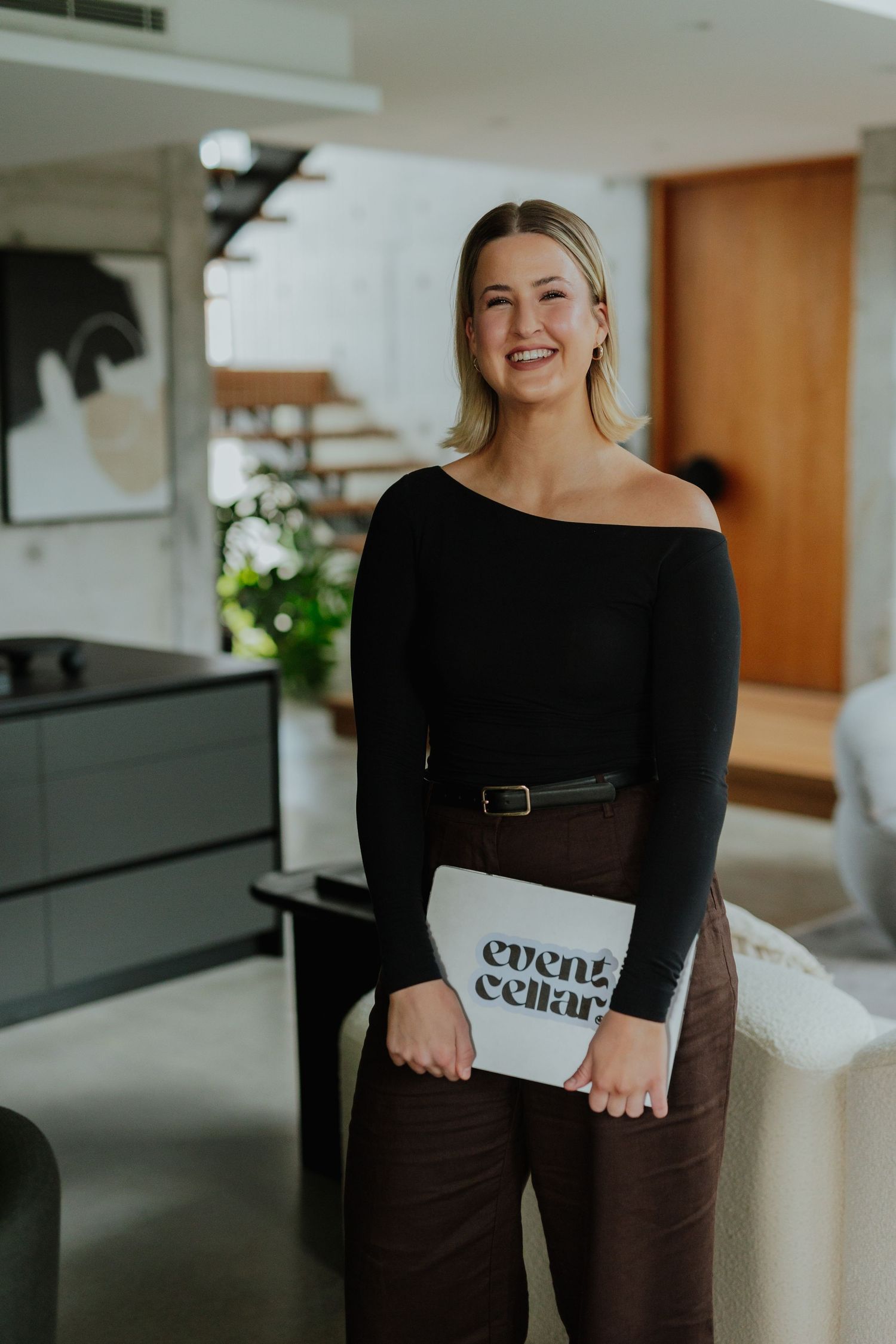 Smiling blonde woman holding an Event Cellar folder wearing a black off-shoulder top and brown pants.
