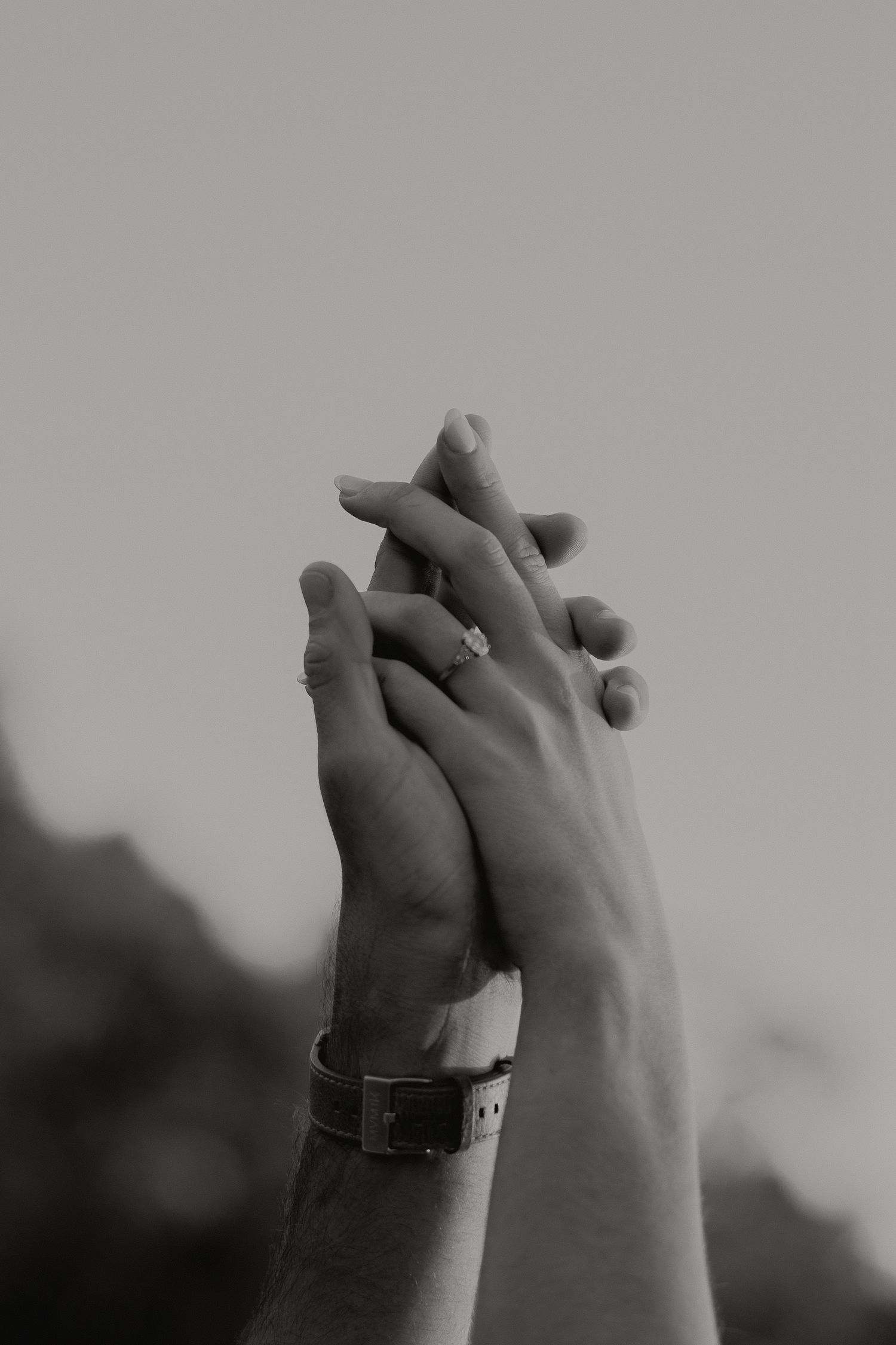 Black and white close-up of two hands intertwined raised upward showing an engagement ring and watch.