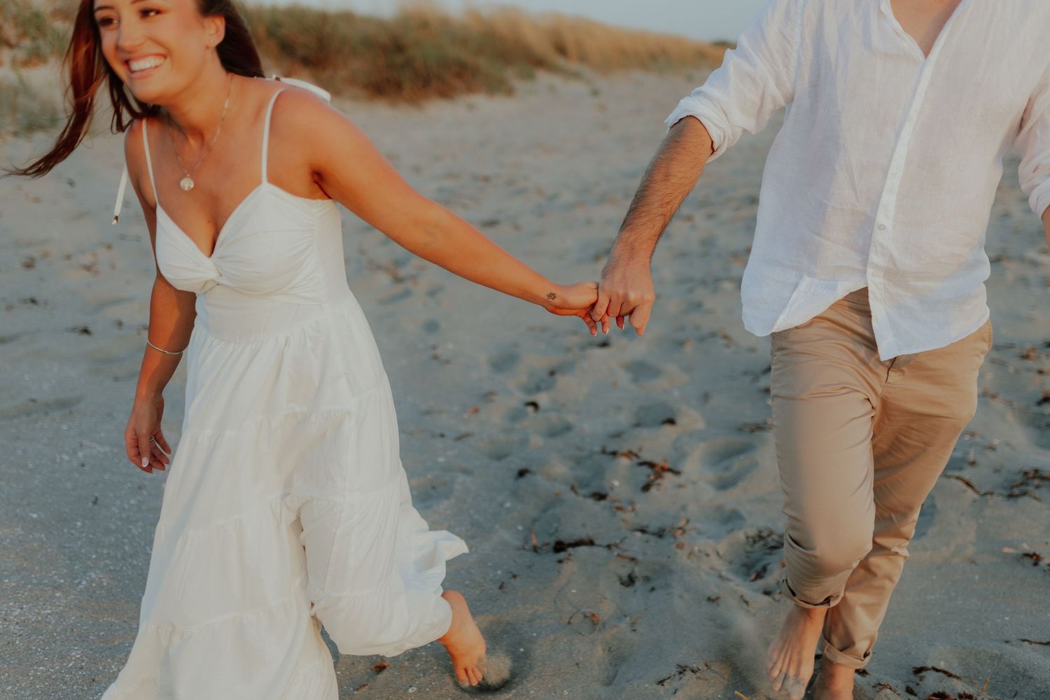 Happy woman in white dress leading man by hand along sandy beach.
