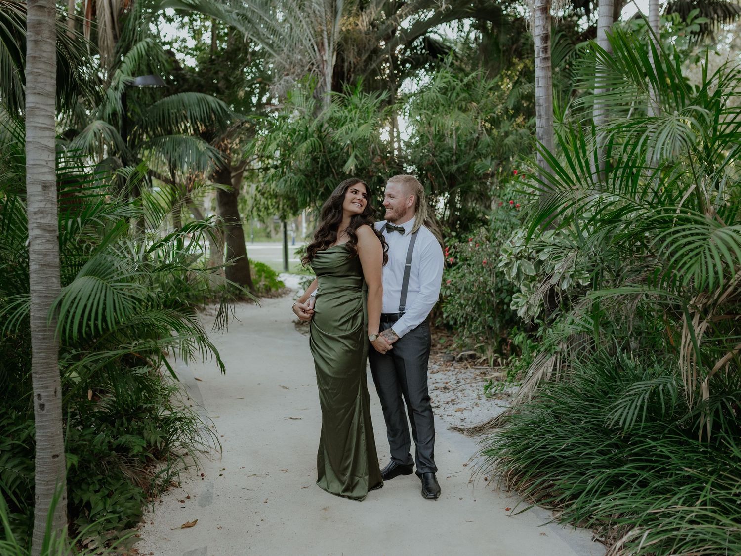 Couple posing on a sandy path surrounded by tropical palm trees, woman in elegant green gown.