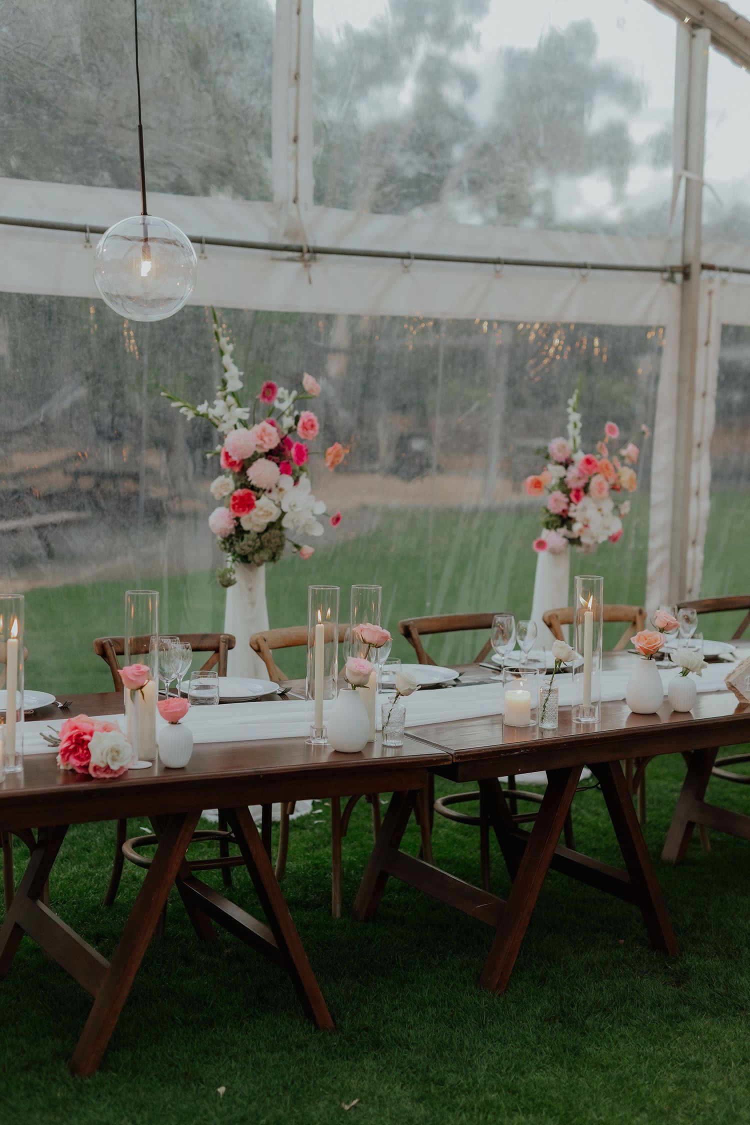 Elegant outdoor wedding table setting with pink floral arrangements, candles, and wooden cross-back chairs under a clear tent.