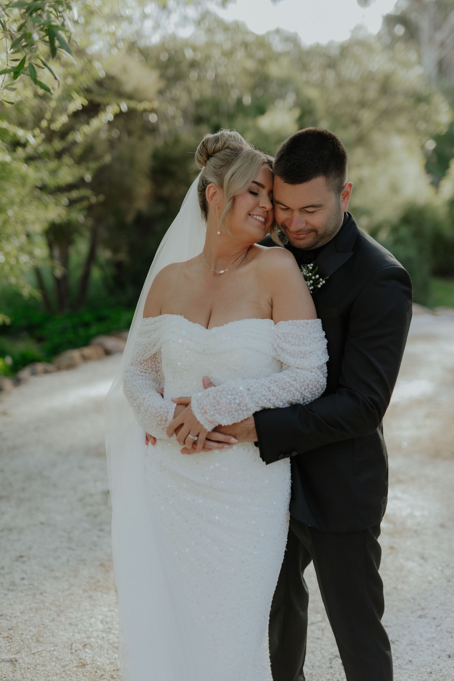 Groom in black suit embraces bride in off-shoulder lace gown on sunlit garden path.