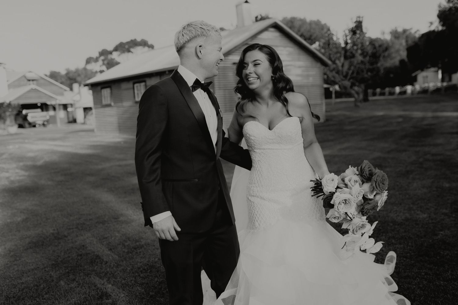 Black and white photo of bride and groom walking together on a lawn outside a rustic venue.