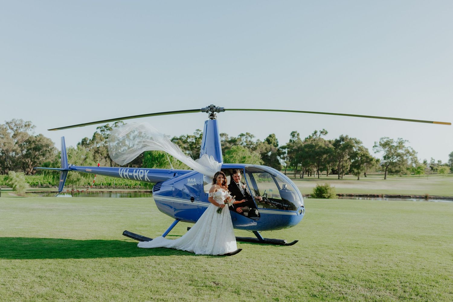Bride and groom posing beside a blue Robinson helicopter on a green lawn on their wedding day.