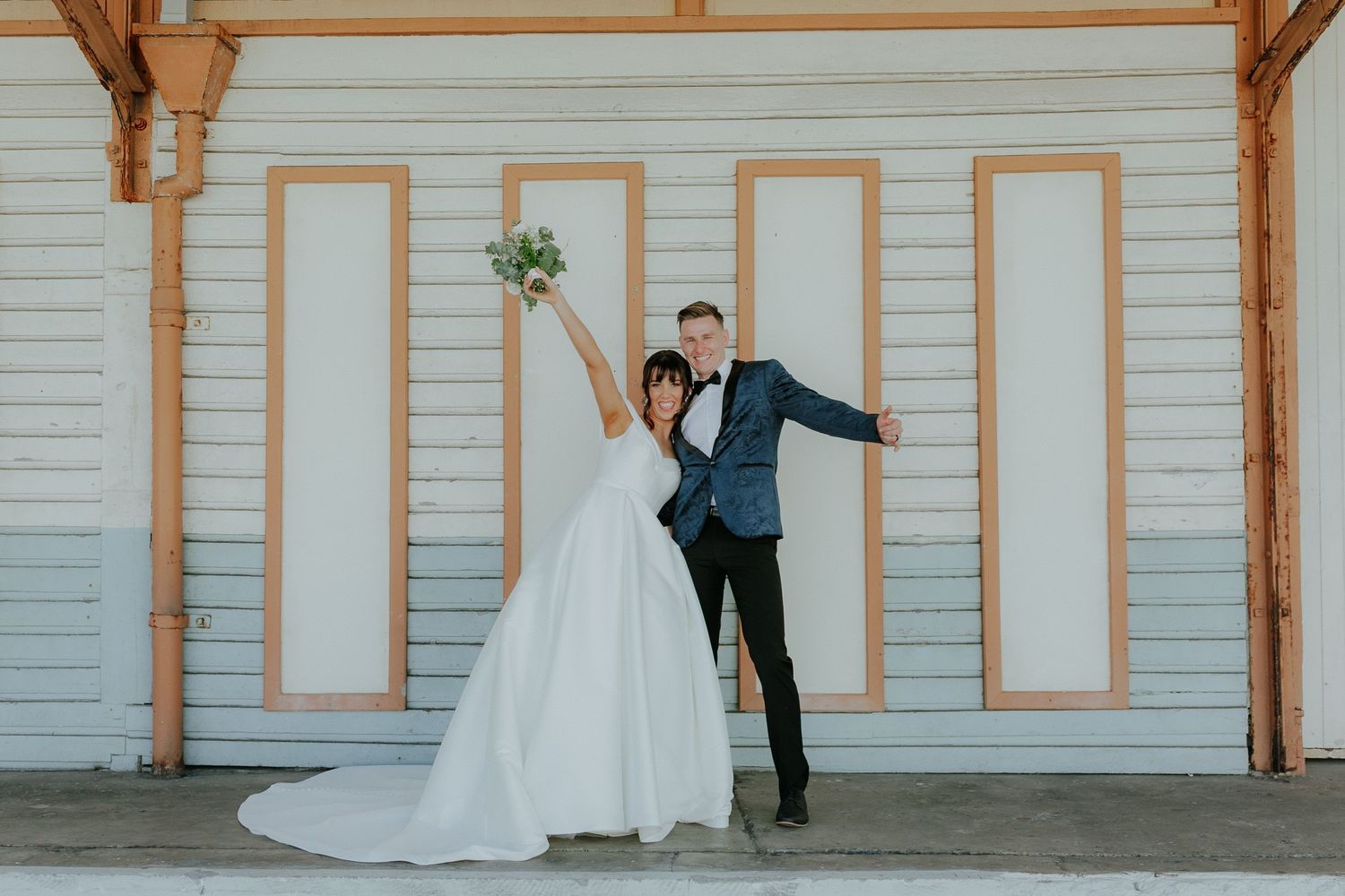 Joyful bride and groom posing in front of white wooden building, bride raising bouquet in celebration.