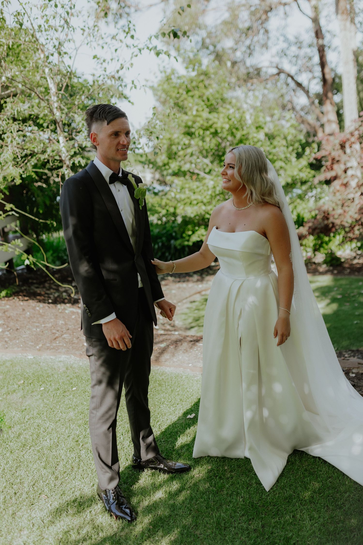 Bride in white strapless ball gown smiles at groom in black tuxedo on lush green garden lawn.