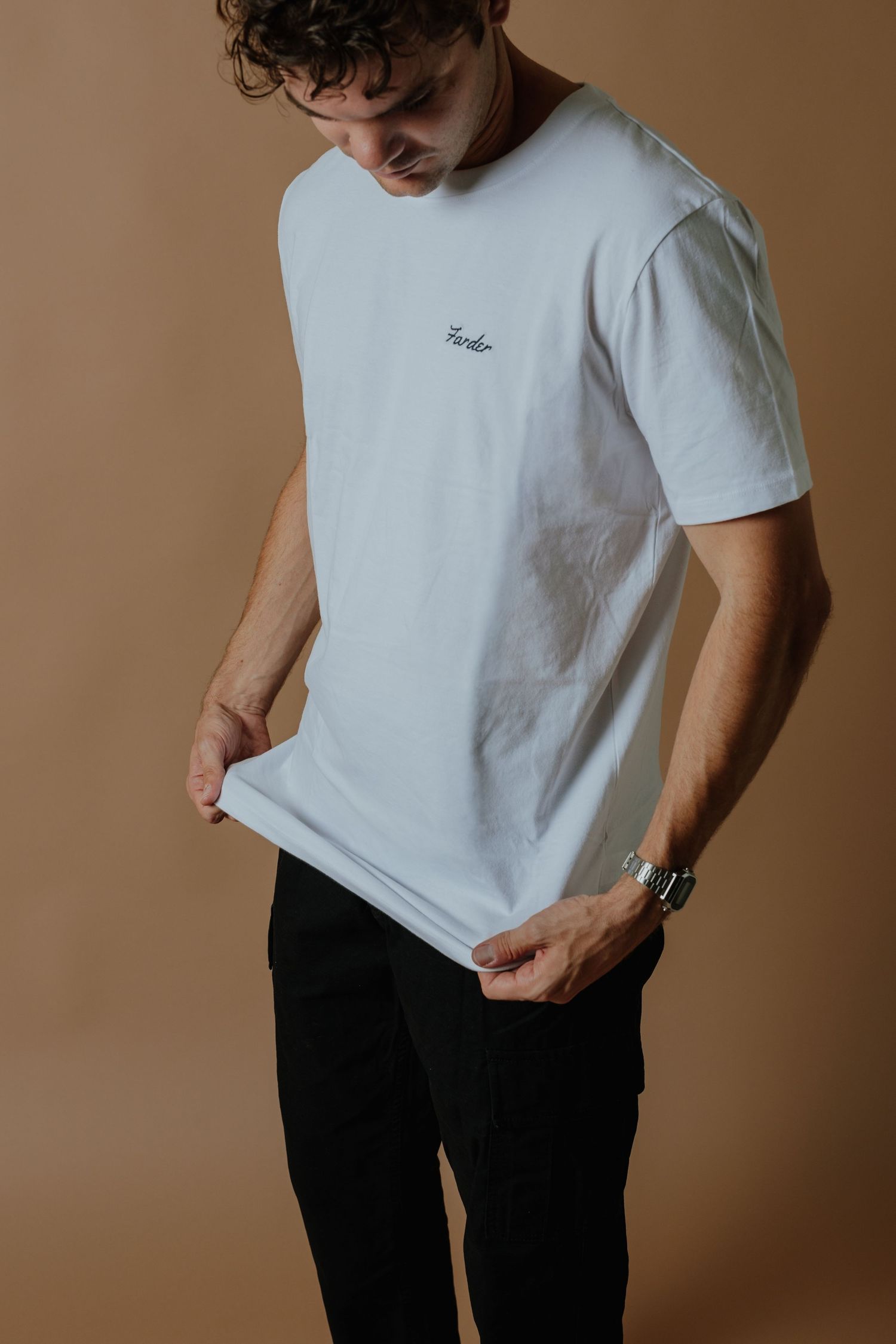 Man wearing a white crew neck t-shirt with script logo, paired with black pants and a silver watch.
