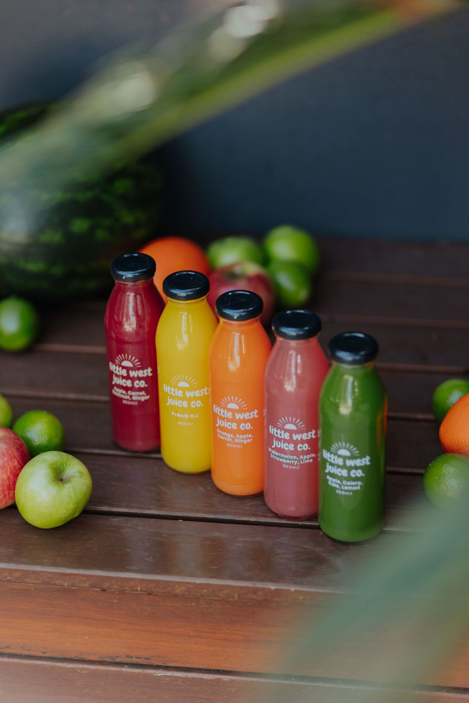 Colorful cold-pressed juice bottles by Little West Juice Co. displayed on a wooden table surrounded by fresh fruits.