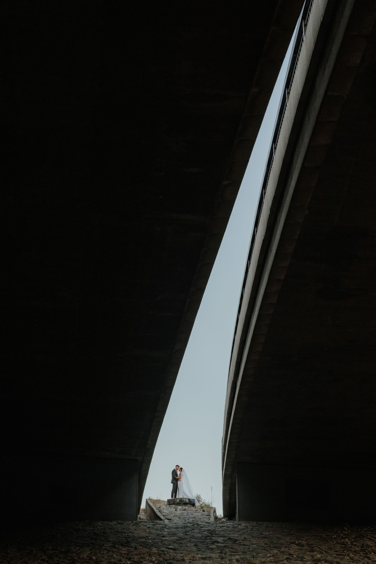 Bride and groom kissing framed dramatically between two concrete bridge structures against the sky.