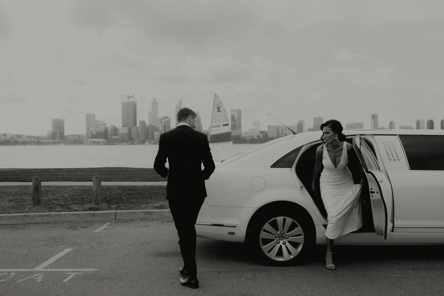Black and white photo of bride exiting a limousine with city skyline visible across the water.