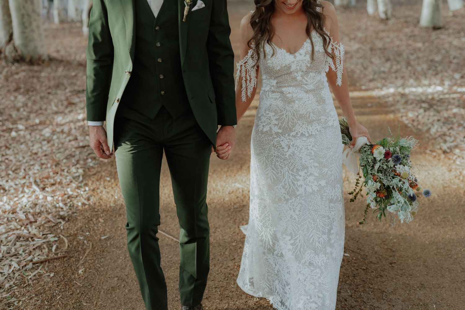 Bride in lace gown and groom in green three-piece suit walk hand in hand through autumn forest path.