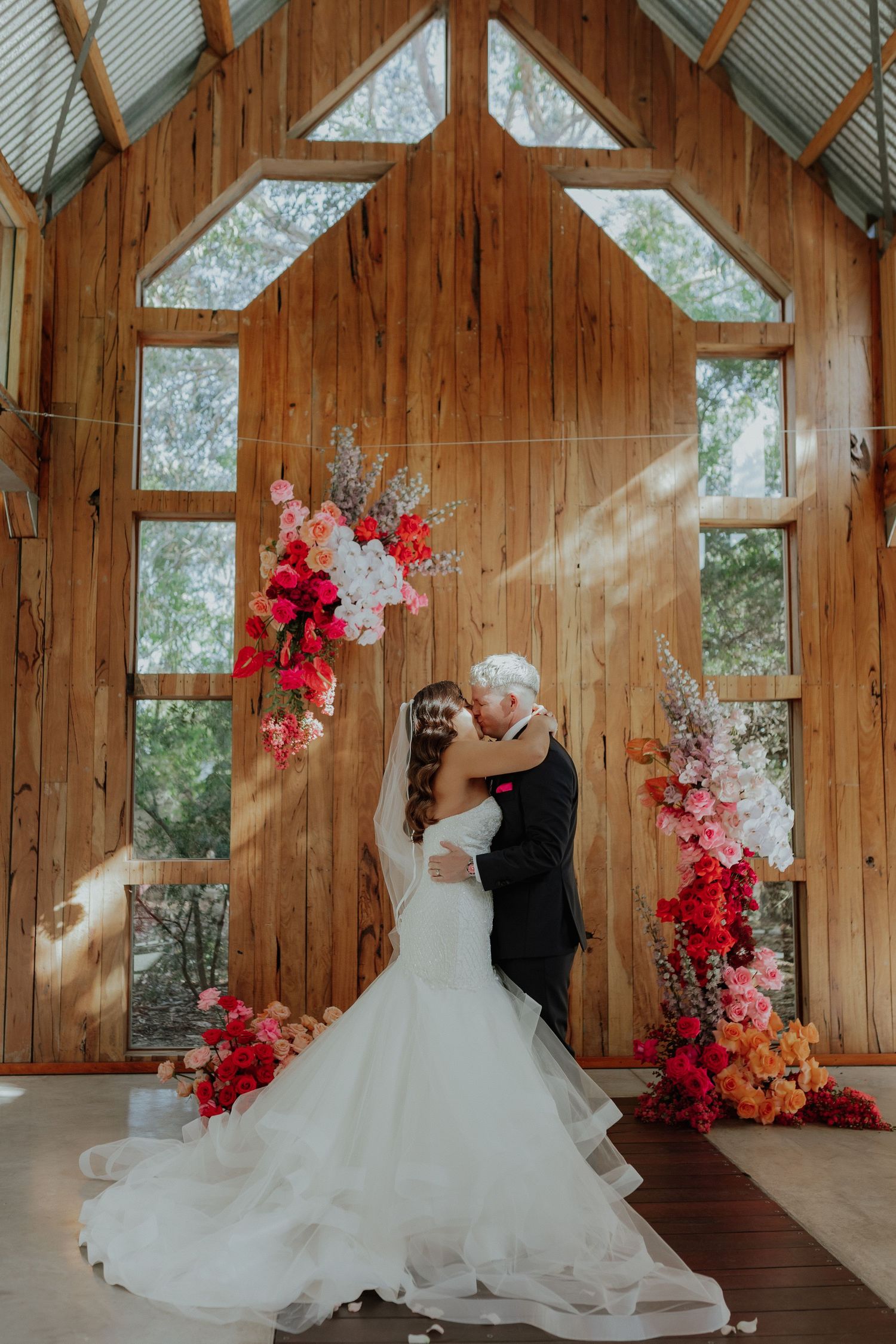 Bride and groom kiss beneath a vibrant pink and red floral arch in a wooden chapel.