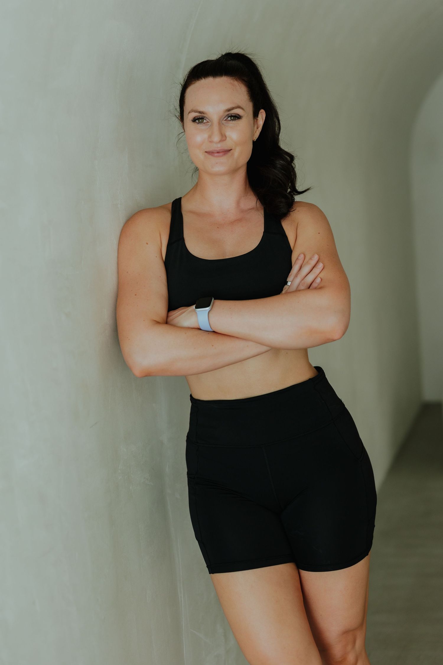 Fit woman in black sports bra and shorts standing with arms crossed against white wall.