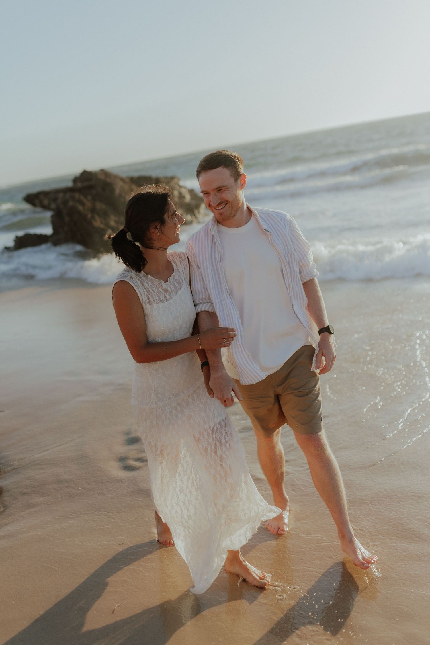Smiling couple walking barefoot on a sandy beach with ocean waves behind them.