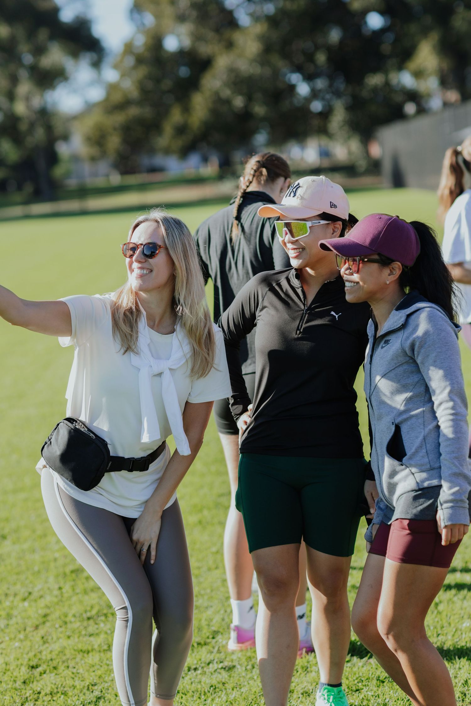 Three women smiling and taking a selfie outdoors on a sunny green sports field.