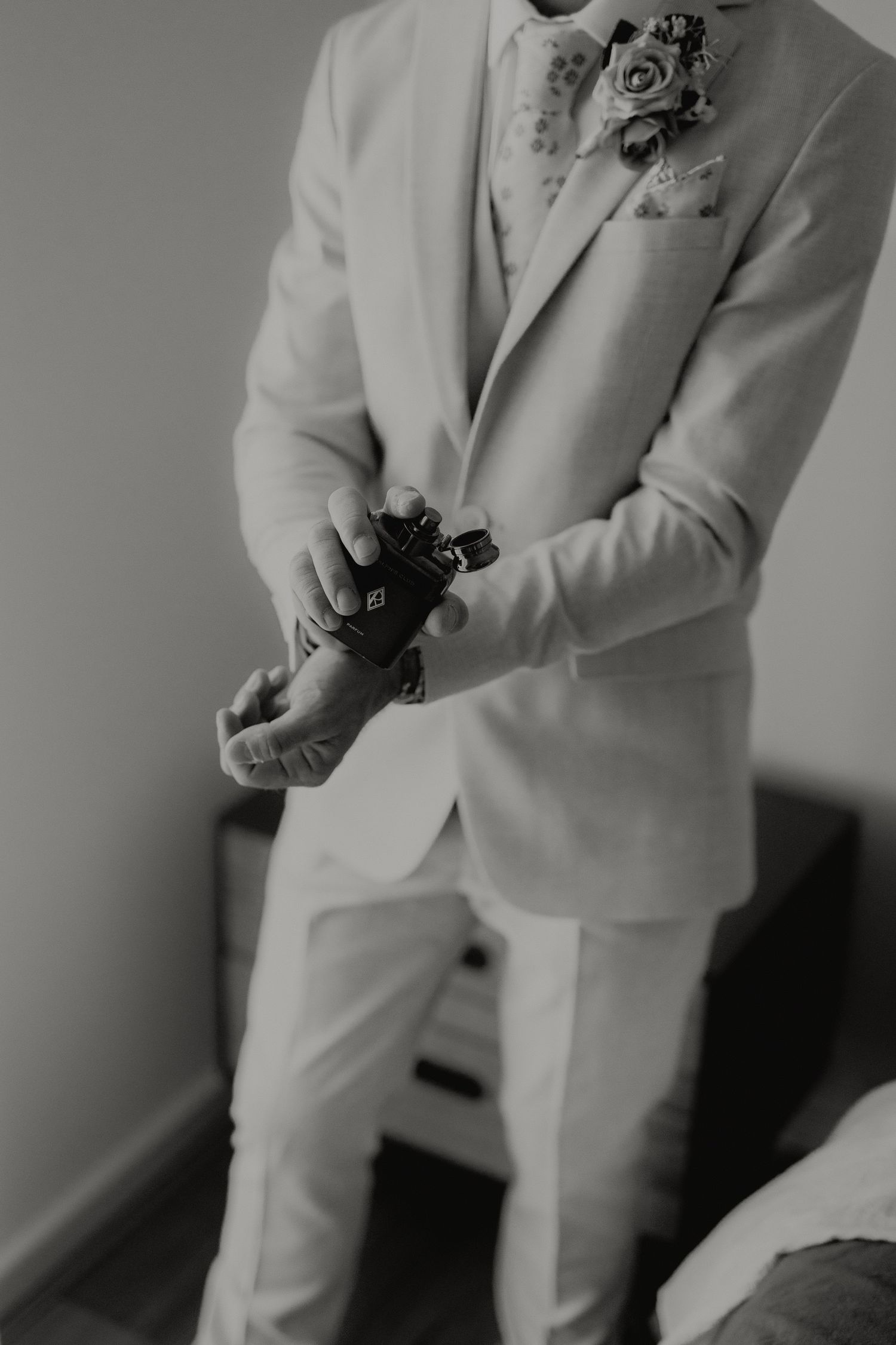 Groom in white suit adjusting cufflinks, boutonniere on lapel, black and white wedding preparation photo.