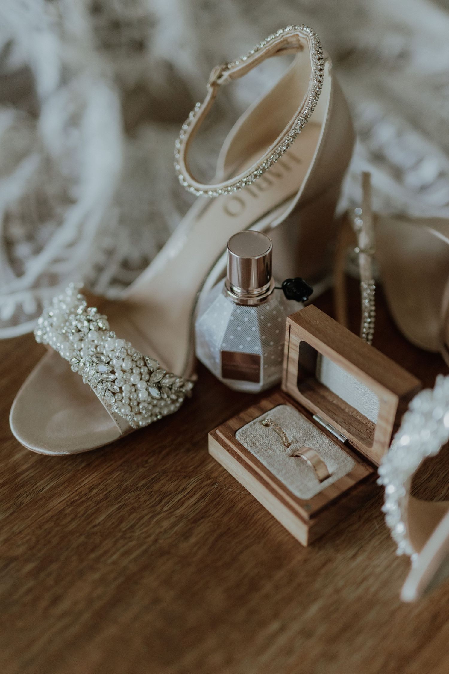 Crystal-embellished bridal heels, wedding rings in wooden box, and perfume on wooden surface.