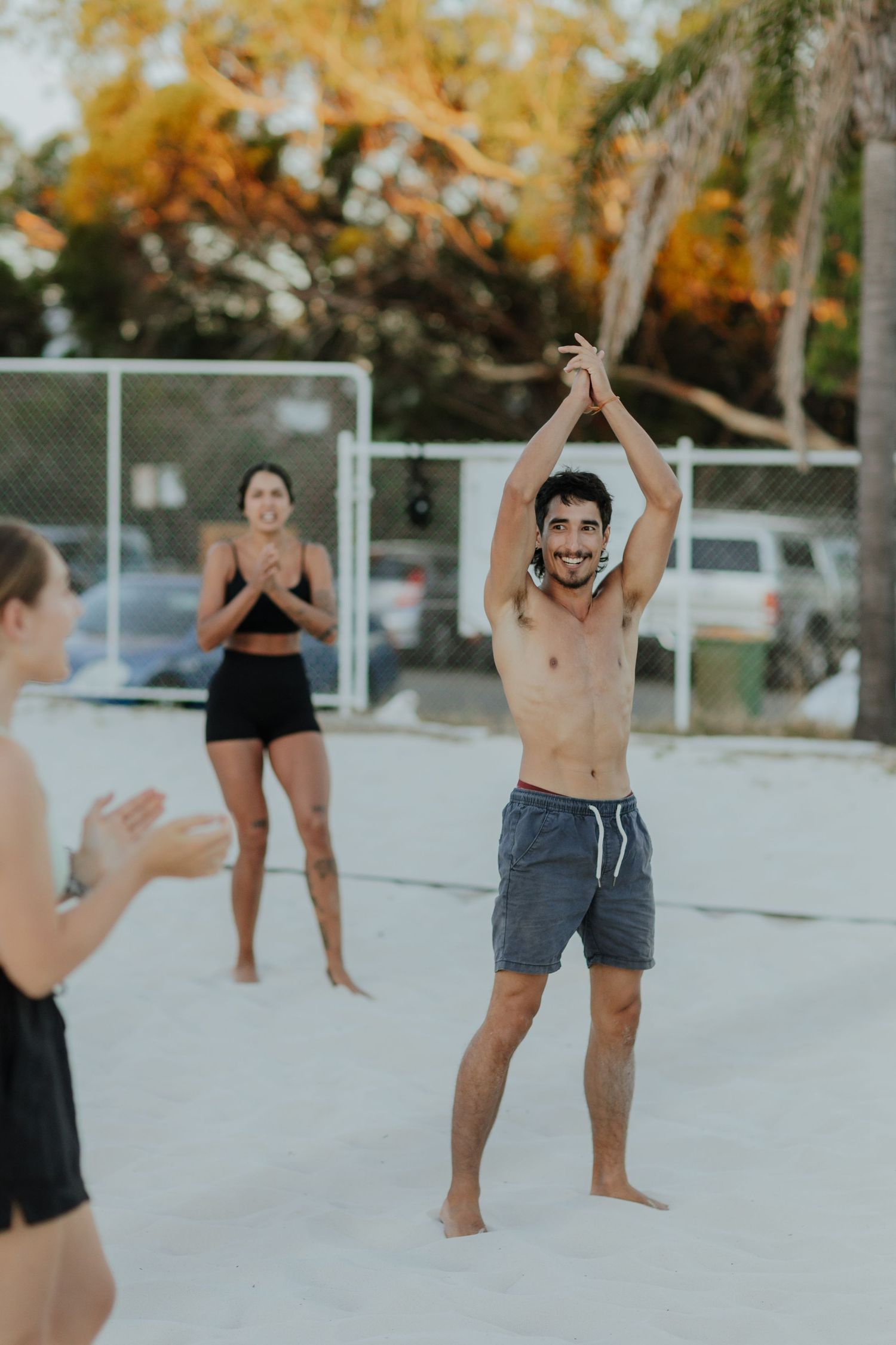 Smiling shirtless man in grey shorts raises hands overhead playing beach volleyball with teammates on white sand.