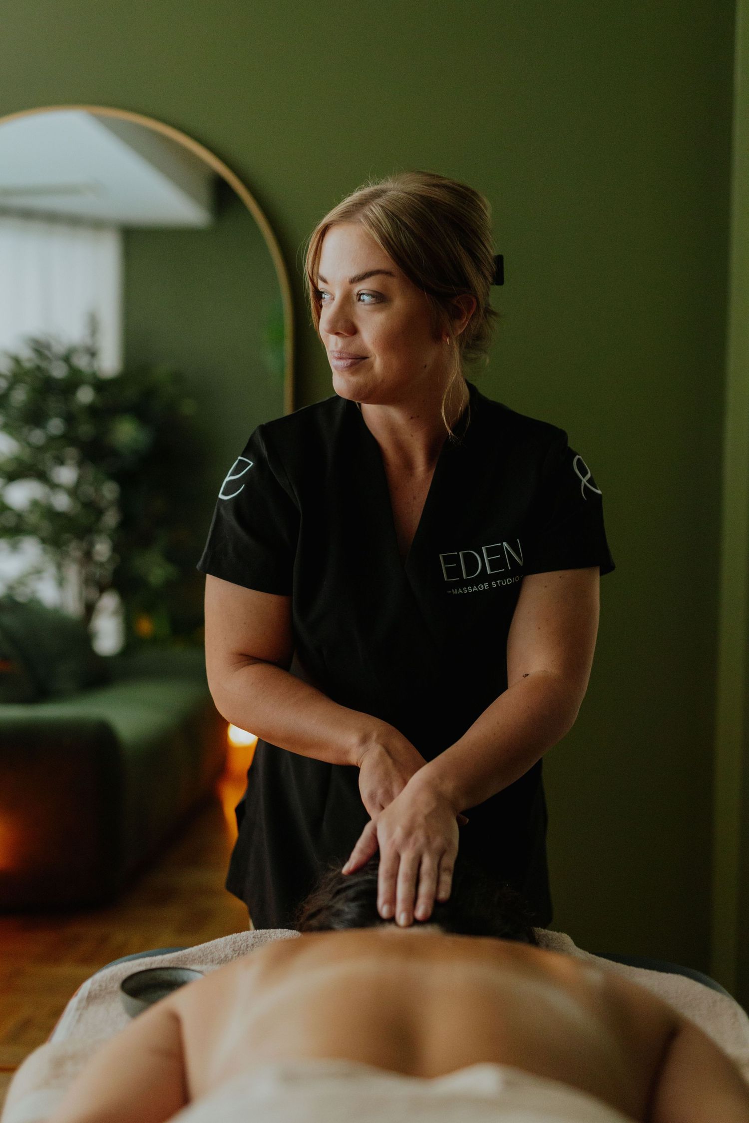 Eden spa therapist in black uniform performing a back massage in a green-accented treatment room.