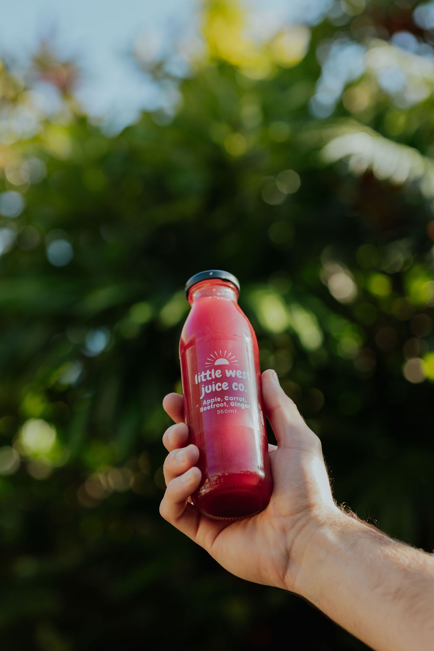 Hand holding a red cold-pressed juice bottle by Little West Juice Co. against a lush green tree background.