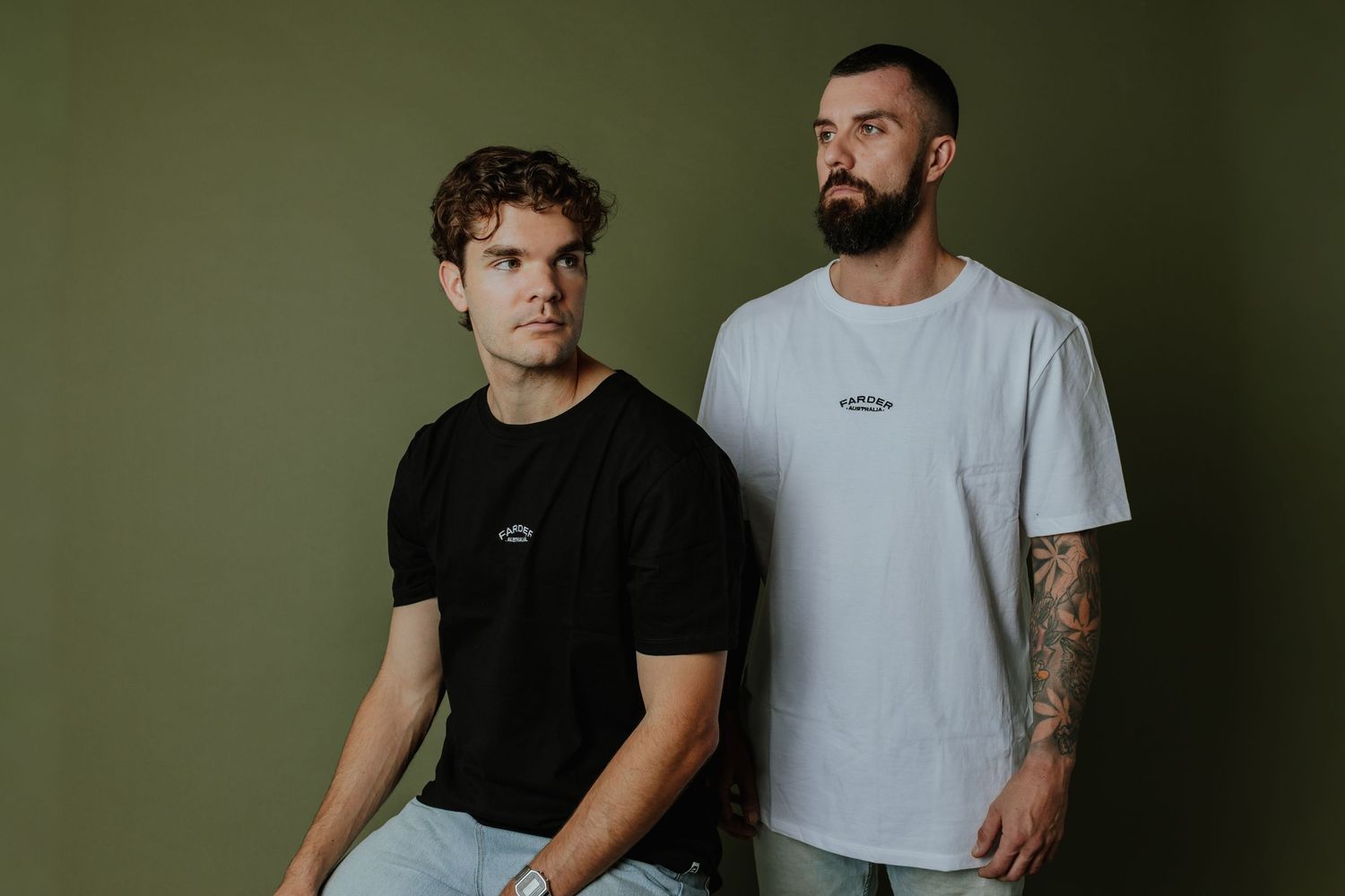Two men posing in black and white branded t-shirts against an olive green background.