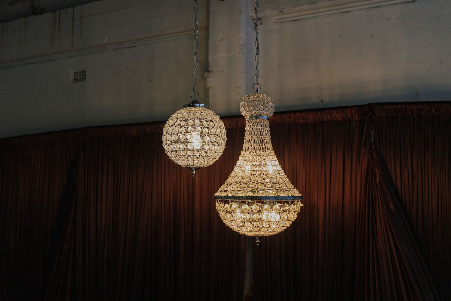 Two elegant crystal chandeliers hanging against dark brown curtains in a dimly lit venue.