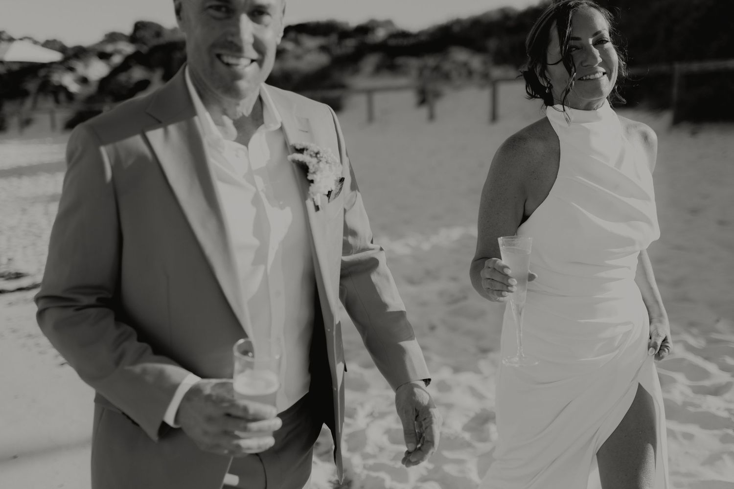 Black and white photo of smiling bride and groom holding champagne glasses on sandy beach.